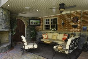 An elegant outdoor patio featuring brick and stone walls, comfortable lounge seating, a ceiling fan, and a wall-mounted TV.