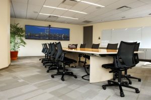 A professional boardroom featuring a long wooden conference table, black mesh chairs, and a large wall-mounted display.