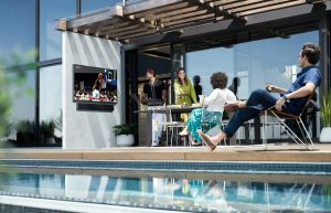 Samsung Terrace outdoor TV and soundbar mounted on a stone wall displaying a basketball game in a poolside lounge area.