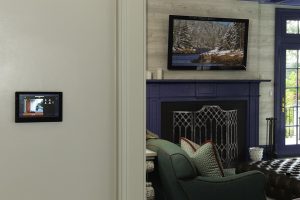 Touch panel mounted in a hallway displaying a camera feed beside an elegant living room with a wall-mounted TV.
