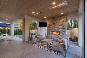 Luxury covered patio with stone fireplace, outdoor TV, recessed lighting, and modern lounge seating at dusk.